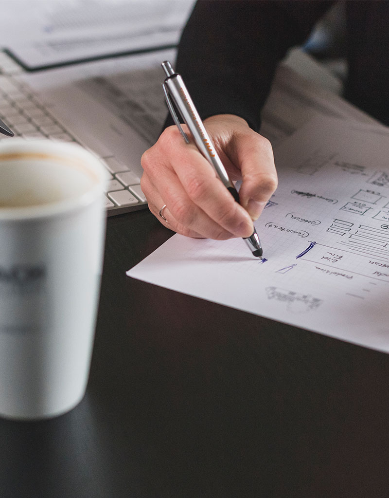 Detailfoto einer Hand mit Kugelschreiber, welche auf einem Zettel Notizen aufschreibt, im Vordergrund ein gefüllter Kaffeebecher