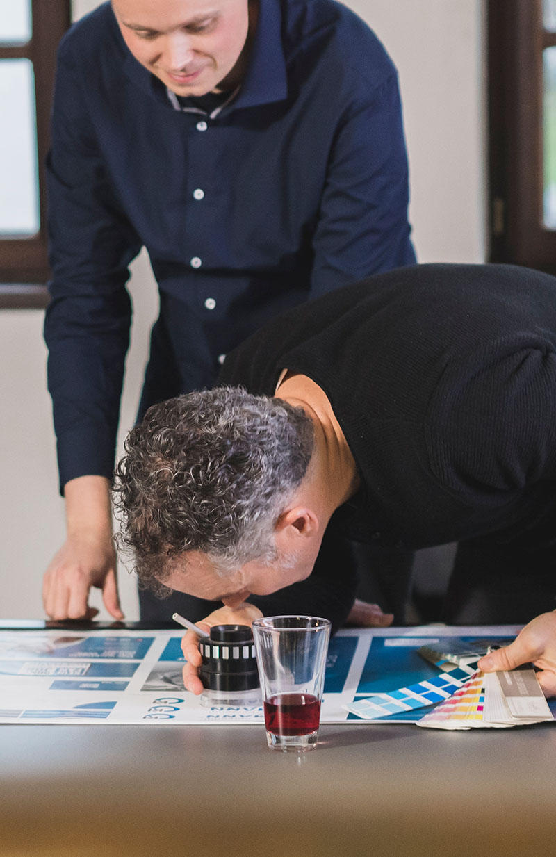 Zwei Personen stehen vor einem Tisch, auf diesem liegt ein Druckbogen, ein Glas mit rotem Saft sowie ein Farbfächer, eine Person ist über eine Lupe auf dem Druckbogen gebeugt
