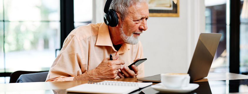 Älterer Mann sitzt vor einem Laptop, in der einen Hand ein Smartphone in der anderen einen Kugelschreiber und blickt angestrengt auf den Bildschirm und trägt Kopfhörer