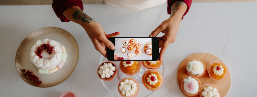 Frau hält ein Smartphone mit geöffneter Kamera und fotografiert Cupcakes und Kuchen