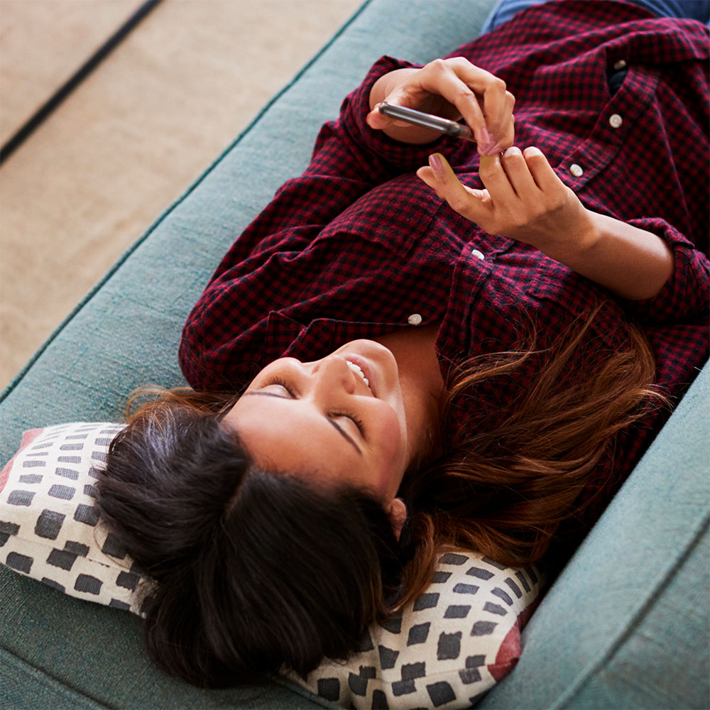 Eine junge Frau liegt auf einem Sofa und hält ihr Smartphone in der Hand, während sie lächelt Eine junge Frau liegt auf einem Sofa und hält ihr Smartphone in der Hand, während sie lächelt