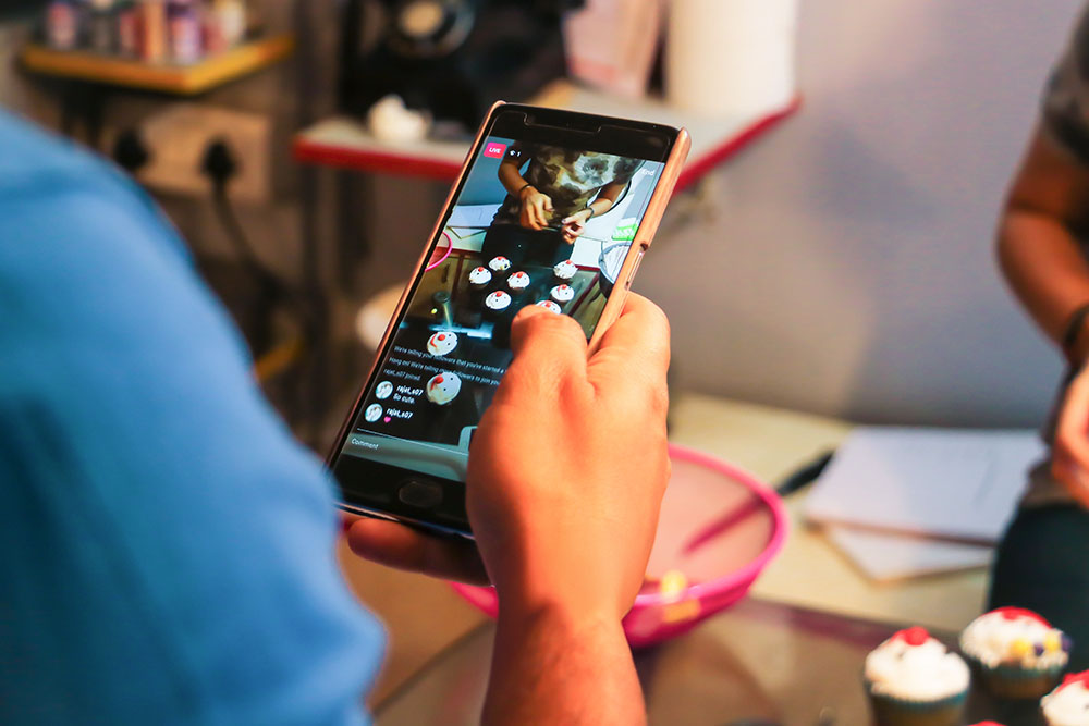 Mann hält ein Smartphone mit geöffnetem TikTok Livestream und filmt Cupcakes