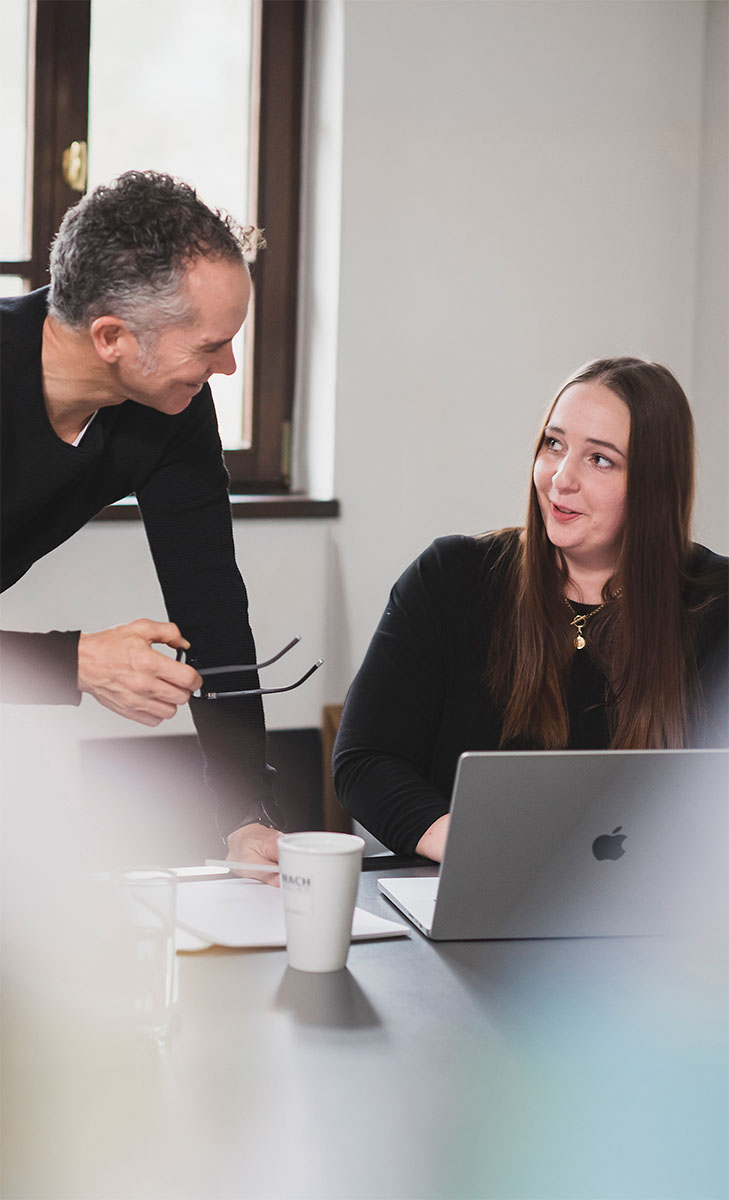Oliver Schneider und Jennifer Reiprich bei einer Besprechung Zwei Personen an einem Tisch, ein Mann steht, eine Frau sitzt vor einem Laptop und beide unterhalten sich