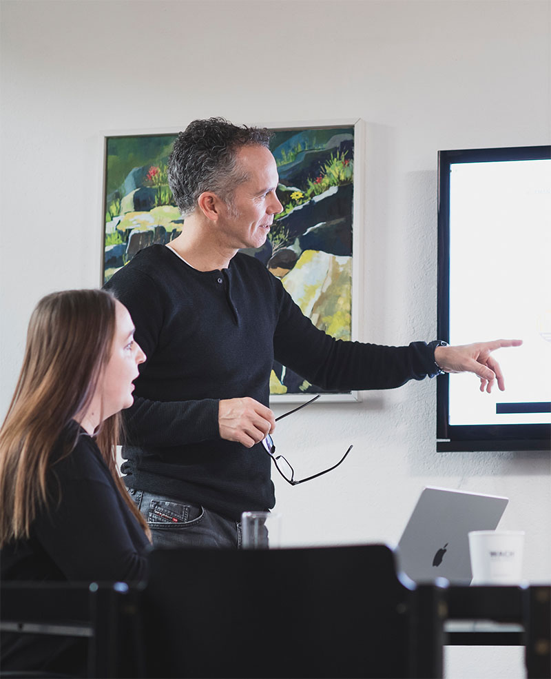 Oliver Schneider und Jennifer Reiprich im Besprechungsraum beim Strategie-Meeting Eine Frau sitzt an einem Besprechungstisch, neben ihr steht ein Mann und zeigt auf deinen an der Wand hängenden Fernseher
